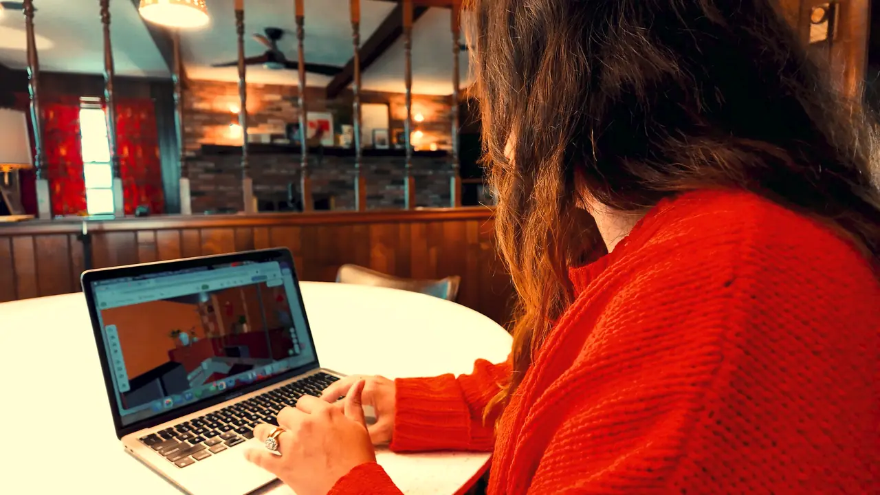 Sarah at a laptop working on a 3D model of a room, a brick fireplace and wood spindles behind her