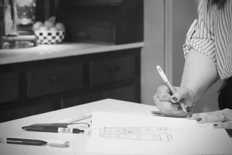 Sarah's hand sketching a room layout at a kitchen counter, in black and white